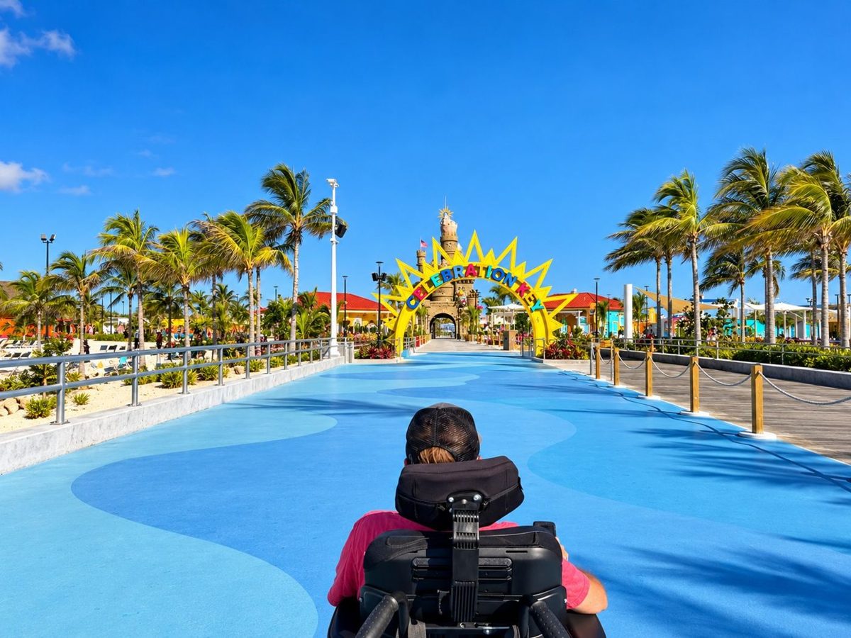 A wheelchair user rolling down a wide, smooth blue path toward the colorful Celebration Key sun arch and sandcastle entrance, lined with palm trees
