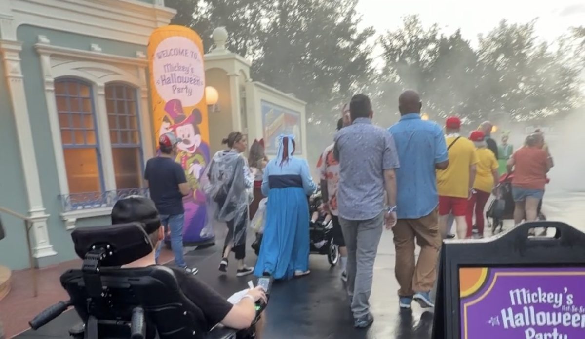 A wheelchair user rolling alongside the crowd past the Welcome to Mickey's Halloween Party banner at the Magic Kingdom entrance