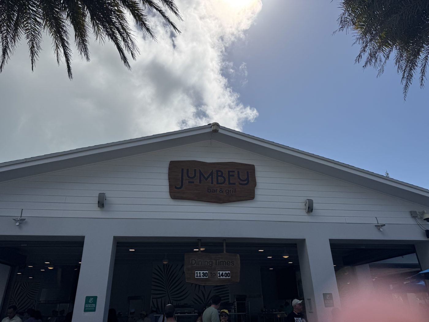 Exterior of Jumbey Bar & Grill at Great Stirrup Cay, with posted dining times and guests gathered beneath the entrance