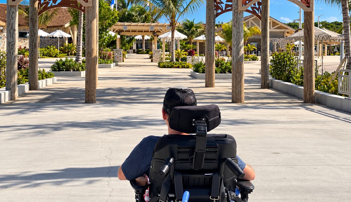 A wheelchair user rolling across a wide, level plaza toward wooden arbors, palm trees and shaded seating at RelaxAway, Half Moon Cay