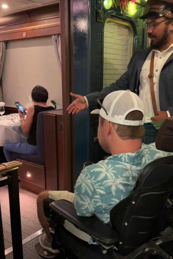 A guest using a power wheelchair being seated by a Royal Railway host inside one of the themed dining train cars
